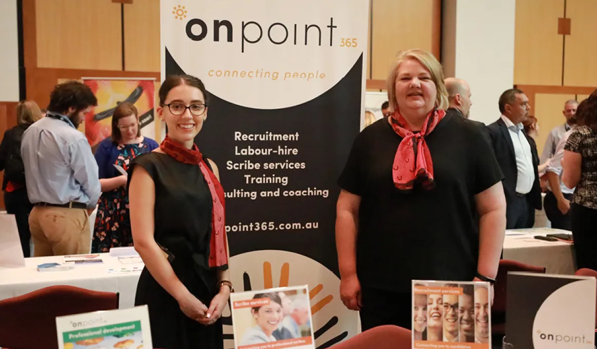 Two women stand in front of a pull up banner that says Onpoint 365, they are facing the camera and smiling. There is a table in front of them and people walking through the background.