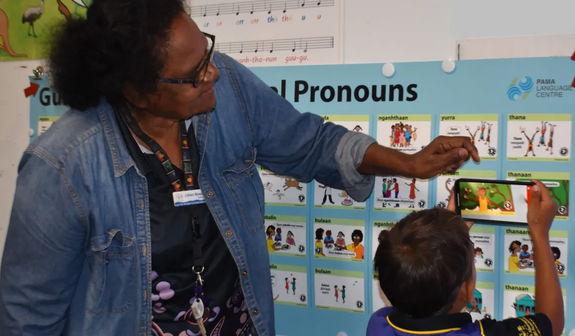 A woman and a young boy stand in front of a poster covered with small images depicting pronouns in language. The young boy holds an iPhone to one of the images to activate the augmented reality. The large poster hangs on a wall along with other posters.