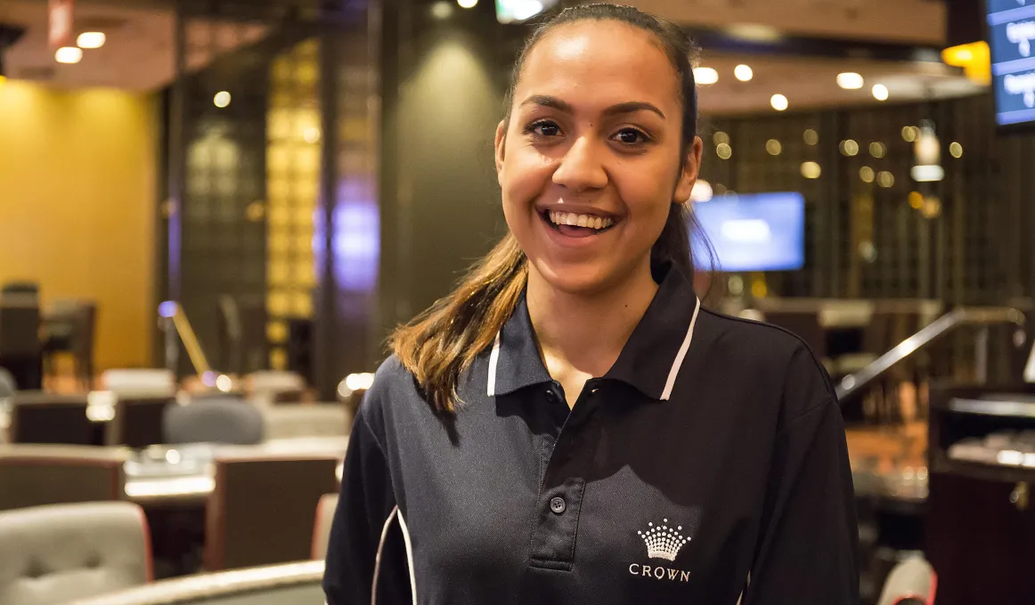 A young Indigenous woman dressed in black polo neck shirt with a Crown insignia stands in a resort lounge featuring lights, booths and chairs.