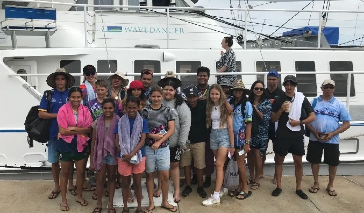 Group of Indigenous Australians, mainly youth, dressed in shorts and casual wear, stand on a wharf in front of a large white boat.