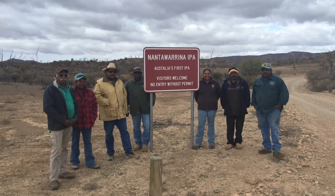 Seven Aboriginal men and women dressed in warm clothing stand next to a red sign just off a dirt road. In the background are bushes, trees and hills. The sign says Nantawarrina IPA – Australia’s first IPA – Visitors welcome – No entry without permit.