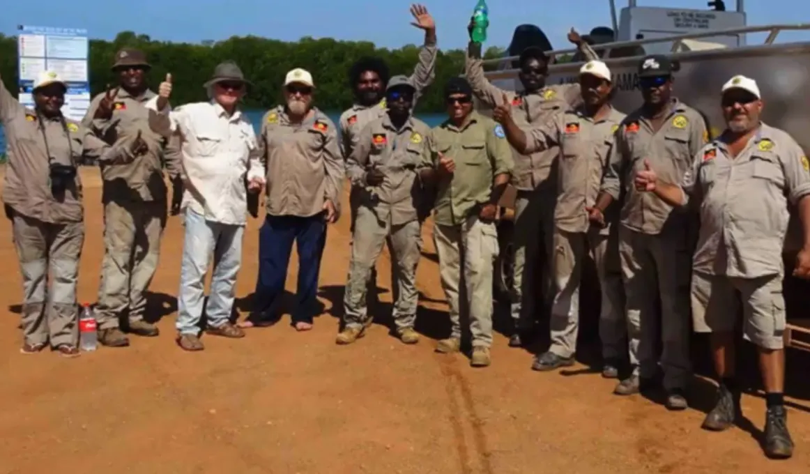 One woman and 10 men, mostly dressed in ranger uniforms, stand on bare ground in front of a river, some signs (cannot read text) and a sliver-grey boat on a trailer.