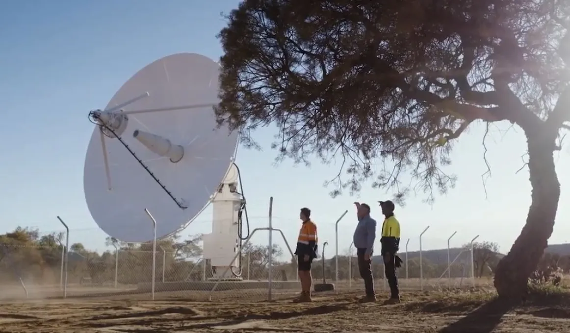 A large and white satellite dish sits on a concrete slab. Three men, two of whom are in work wear look at it. At right is a tree and in the background are hills and vegetation.