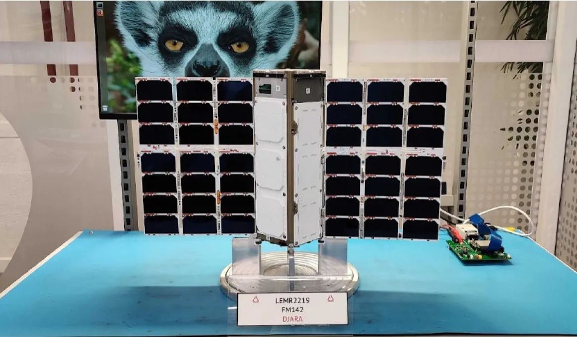 A mechanical device with white panelling and banks of black coloured photovoltaic cells on either side sits on a blue table. Behind, on the table, is an electronic device and in the background is a wall including a picture of a Lemur.