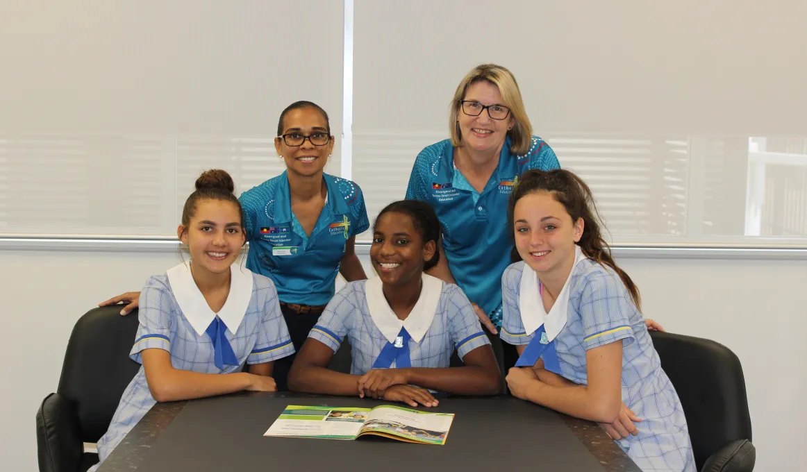 Two women in aqua coloured shirts stand behind three young women in pale blue school uniforms seated at a table.