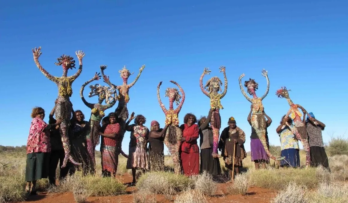 Thirteen Indigenous women stand in a line. They are holding up 7 life sized artistic figures of women.