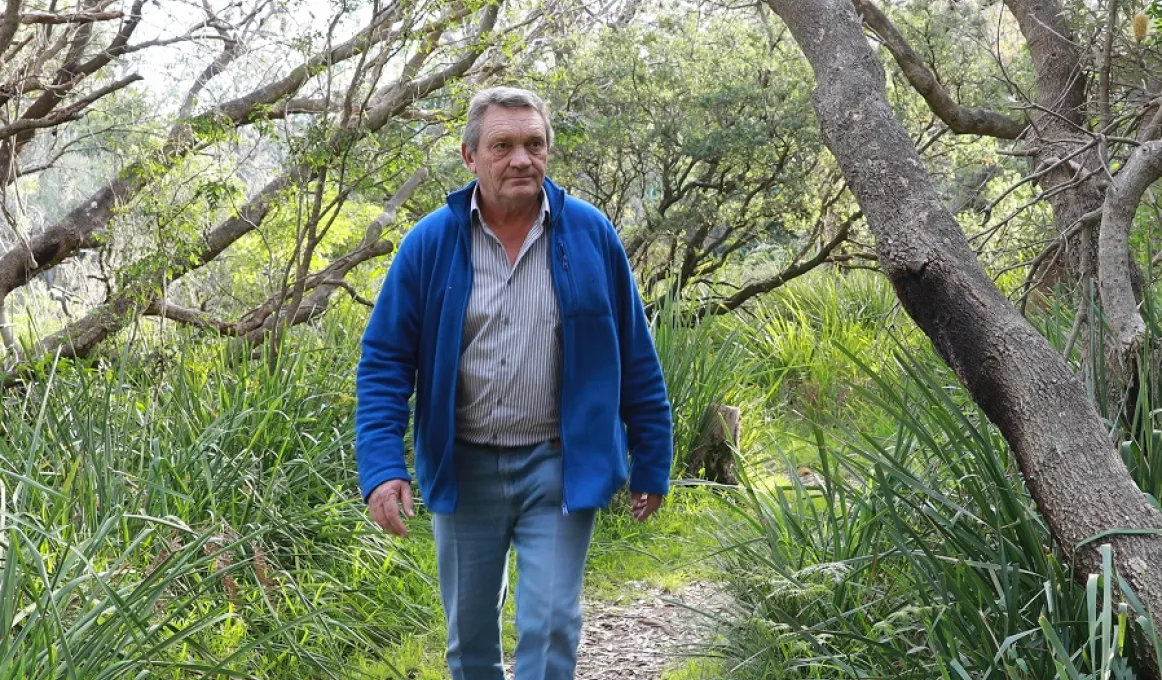 A man in jeans and a blue jacket walks along a bush path, with trees on both sides of him and native grass at his feet.