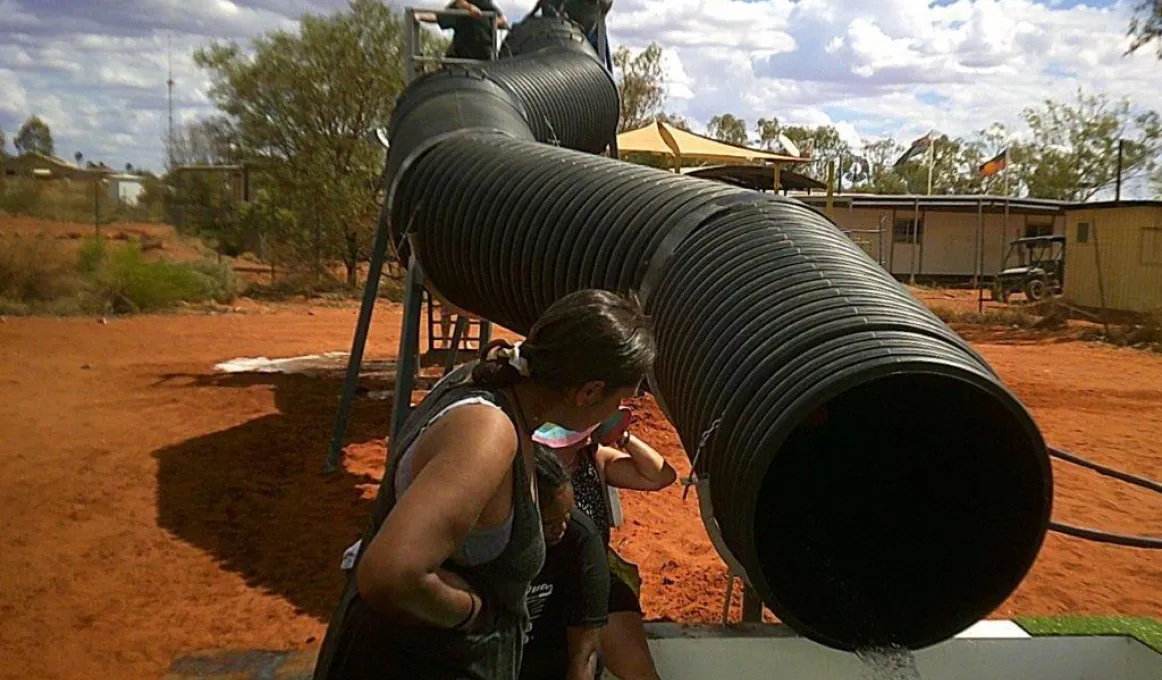 A long and very wide black pipe snakes from a stand downward to a pool. On the stand are several people and at the pool are a few more. In the background is red soil, trees and buildings.