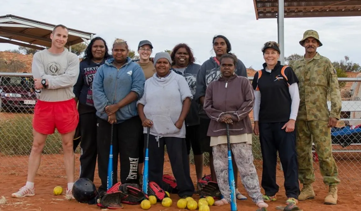 Indigenous women with non-Indigenous army men and women wearing various clothing types and some holding softball bats. There are softballs on the red earth in front of them.