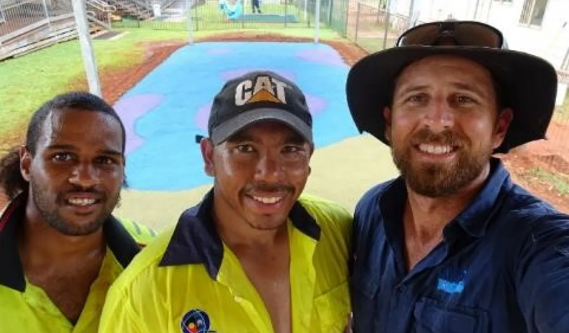 Three men, two in yellow work-wear shirts and one in a blue shirt stand close to camera. In the background is a flat area covered with blue and pink rubber. Surrounding this is bare soil, grass and fences. There are buildings at right and left and behind.