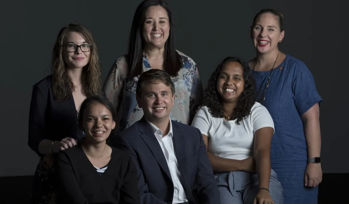 Group of 5 Indigenous women and one Indigenous man smiling to camera in front of dark background.