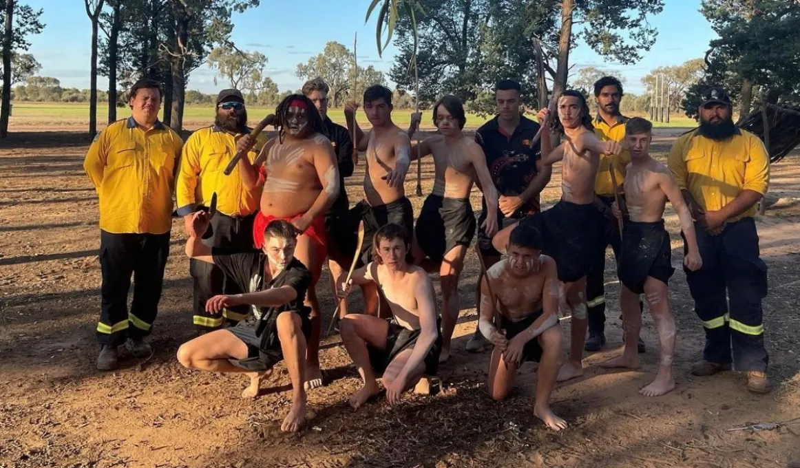 Group of young men dressed in traditional wear and paint stand or kneel on dry ground. With them are men in firefighting wear. In the background are trees, a grassy field and a blue sky.