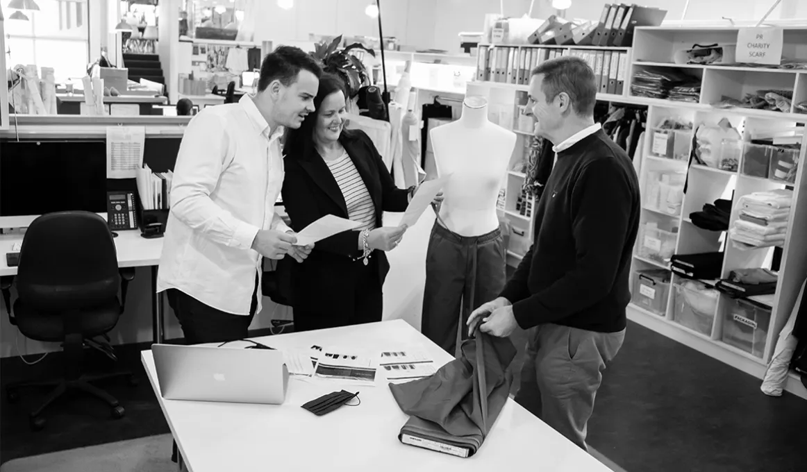 Two men and a woman in casual clothing stand next to a table on which is a laptop, some papers and a roll of material. In the background is a desk and shelves filled with papers, boxes and office supplies.