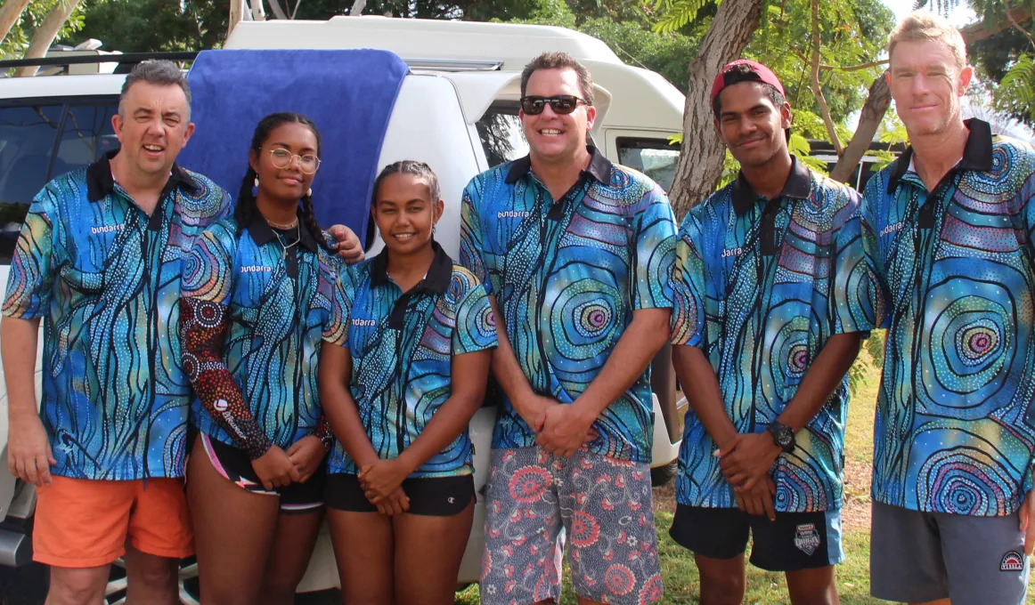 6 adults (2 female and 4 male / 3 Aboriginal) dressed in blue shirts featuring Indigenous designs stand in front of a white car on grass with trees in the background.