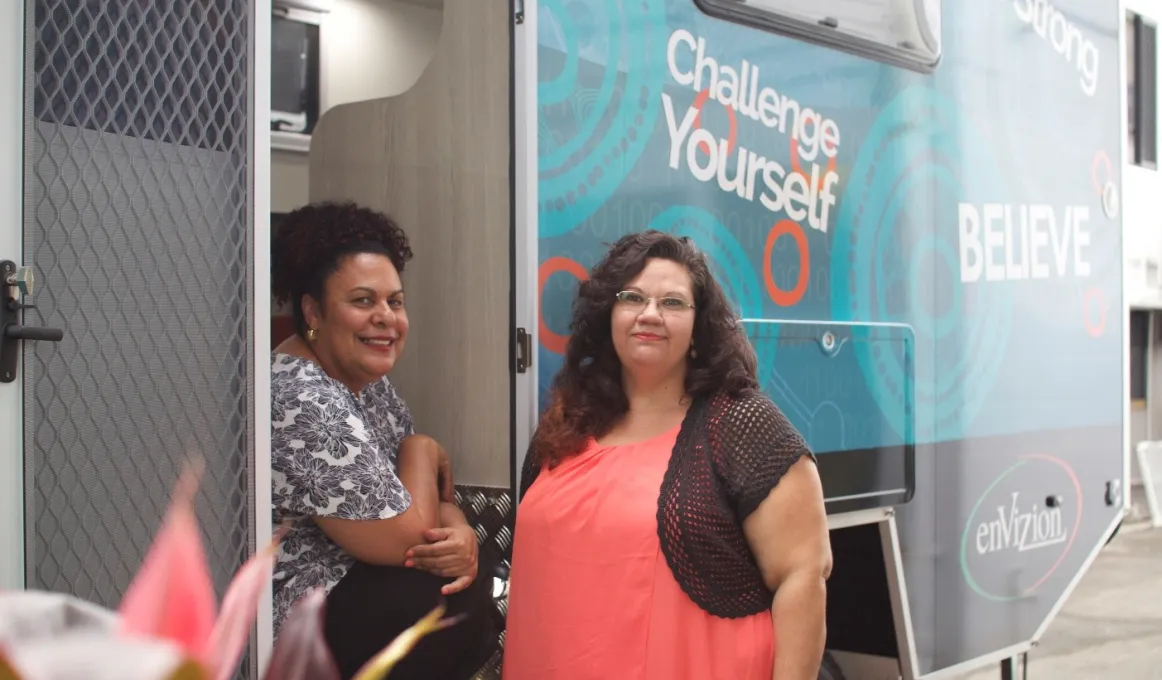 An Indigenous woman in grey and white top sits on steps of bus while another Indigenous woman in apricot dress stands next to her. Words on the bus include Challenge Yourself, Believe, enVizion and Be Strong.