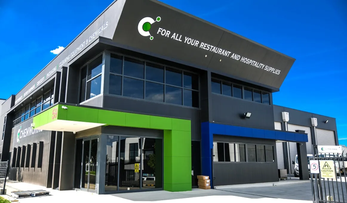 Image is a modern black building with windows and a green entrance. At right are large roller doors. The entire foreground is a concrete slab or driveway.