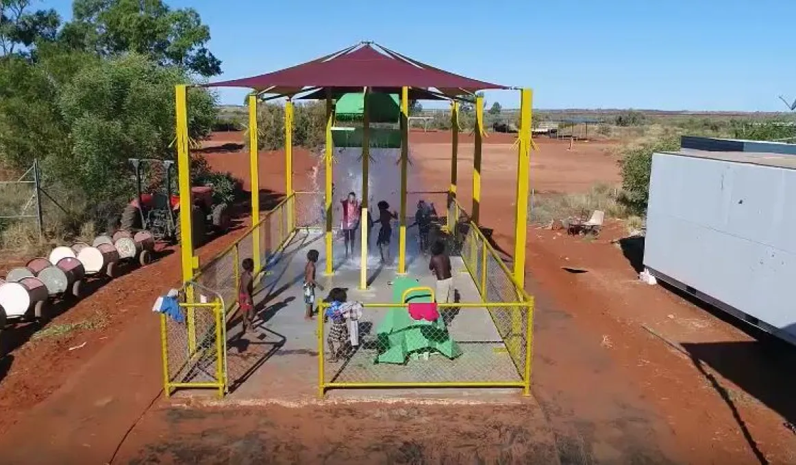 Several Aboriginal children play on a concrete slab under sun shade amongst various water features. On the left is a small train with carriages made from barrels and on the right is a building. In the distance is open ground, more structures and bushland