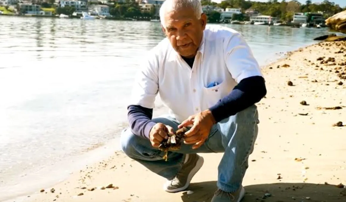 Elderly Aboriginal man in white shirt and jeans squats next to a river on a sandy shore. In the background on the other side of the river are houses and trees.