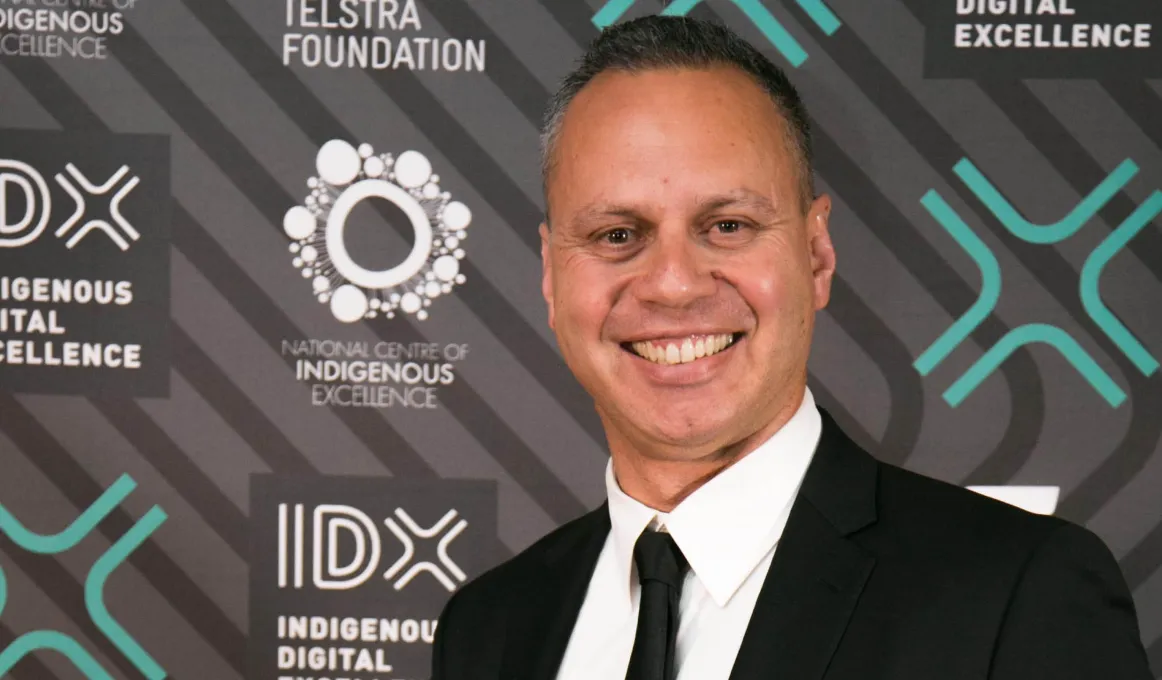 Indigenous man in a black suit and white shirt standing in front of a backdrop featuring various logos including IDX Indigenous Digital Excellence, Telstra Foundation and National Centre of Indigenous Excellence.