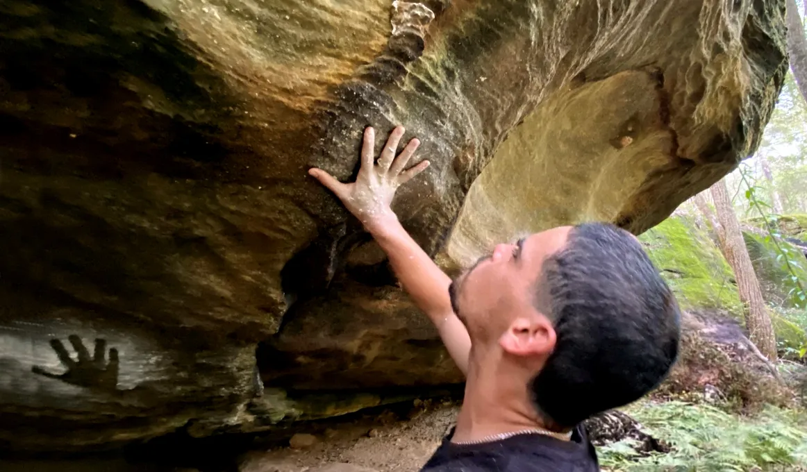 A young adult man stands beneath a rock overhang and touches the rock with his hand. On the rock is another hand print. In the background are more rocks and trees.