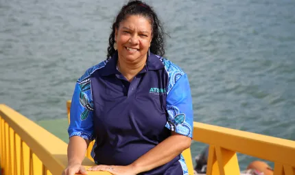 Aboriginal woman in blue shirt stands next to a yellow rail with the ocean in the background.