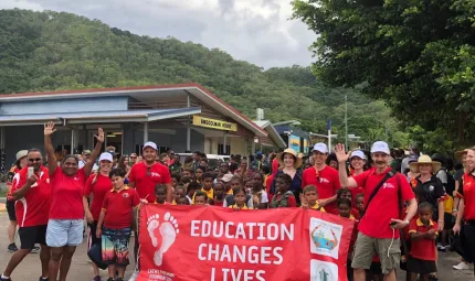 A group of men, women and children in red school polo shirt with a crowd of Indigenous children behind red banner with white writing on banner ‘Education Changes Lives.’