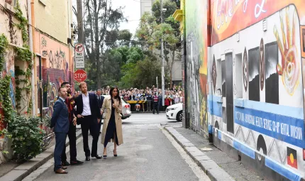 4 people (Duke and Duchess of Sussex, an aboriginal man Robert Young and third man) stand in an alley viewing colourful murals on the walls lining the alley. In the background are cars, a crowd of people, and trees behind the crowd.