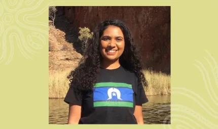 Indigenous woman with long black hair wearing a black t-shirt with a blue, green and white flag on the front stands in front of body of water. In the background is grass and a cliff face.