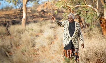 A woman wearing a black top, orange floral skirt and leopard print cardigan is walking in the scrub surrounded by trees. The woman is indicating to something in the distance that is not visible in the image.