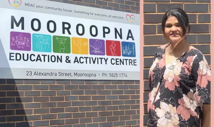 At left is building wall with a large sign featuring the words: Mooroopna Education and Activity Centre. At right is a woman with black hair and wearing a dark dress with large white, yellow and pink flowers.