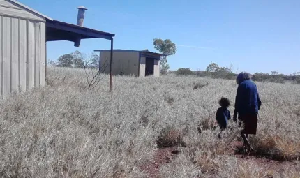 In the grassy scrub there is an old woman in a blue cardigan walking with a young child in a blue t-shirt towards some buildings.