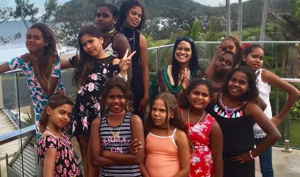 Aboriginal woman with a group of 12 year 5 and 6 Aboriginal girls. They are wearing colourful dresses, and a standing on a balcony with a beach behind them.