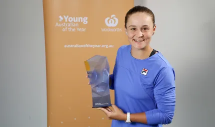 A young woman holding a glass trophy wearing a blue shirt is standing in front of a yellow screen that reads “Young Australian of the Year”.