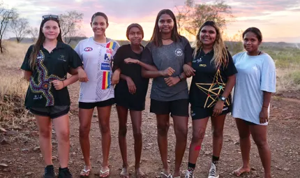 Six young Aboriginal women stand arm in arm on bare soil with dry grass, a few trees and a setting sun in the background.