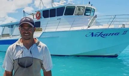 A man stands at the front of a small boat dressed in casual wear including a cap. In the background is pale blue water, a mainly blue sky and a large boat with a cabin and the words Nikana II painted on its front.