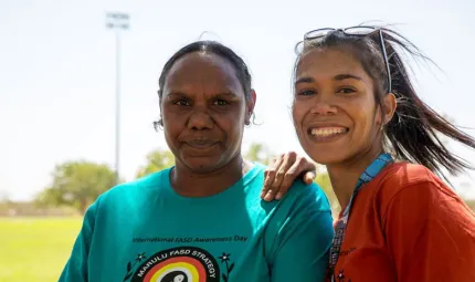 Two women stand shoulder to shoulder. The woman on the right in an orange shirt has her hand on the shoulder of the other who wears an aqua coloured shirt.