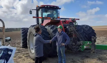 A woman in a long grey coat shakes hands with a man in jeans and top. In the background is a tractor and open field.