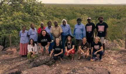 A group of people dressed in casual or work wear stand or squat on stony soil near the edge of a drop off. In the background is a tree covered plain and cloudy sky.