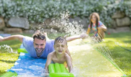 A small child holding a green inflatable doing down a slip ‘n’ slide in the back yard. Behind his is an older man on the slip and slide. In the background is a young girl holding the hose on the waterslide