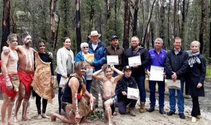 11 people standing and three kneeling on sand in front of trees. Most of the people are holding paper and some are wearing Wiradjuri traditional dress.