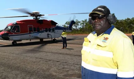 A man in work wear and including safety headphones and a cap stands on an aircraft tarmac. In the background are other men and a large helicopter. Behind that is bush land.