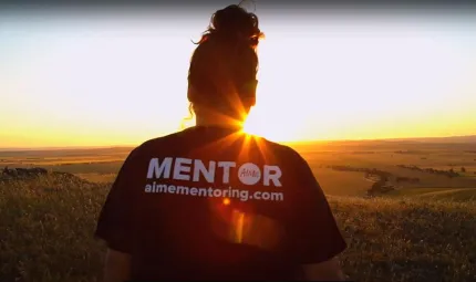 A woman with long hair tied up in a bun looks out on a landscape and the sun as it sits just above the horizon. She wears a t-shirt with text on the back. It says Mentor aimementoring.com