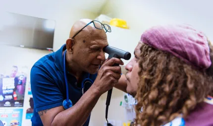 Male doctor in blue shirt uses an instrument to check the eye of another man with dreadlocks and wearing a pink beret. In the background are posters.