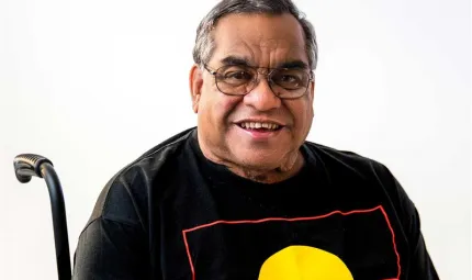 Close up image of a middle aged man sitting in a wheel chair. The man is smiling and wearing glasses and a t-shirt with the Aboriginal flag on it.