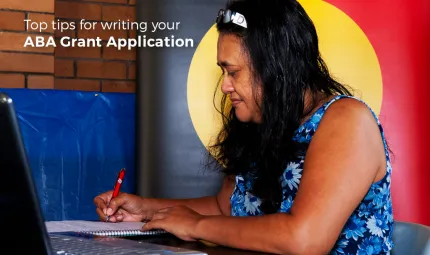 Indigenous woman writing on paper with a pen and the Aboriginal flag in the background, a laptop computer in the foreground and the following words displayed: Top tips for writing your Aboriginal Benefit Grant Application