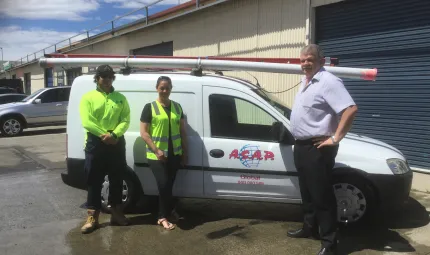 An Indigenous young man and young woman wearing safety tops and an older man dressed in light shirt and dark trousers stand in front of small white van parked in front of yellow building with dark green roller door. Letters on the car door are A.S.A.P.