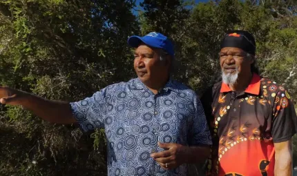 Two Aboriginal men stand side by side. The man on the left points to his right. He wears a blue and white shirt and blue head covering. The man on the right wears a red, brown and yellow shirt and similar coloured head covering.