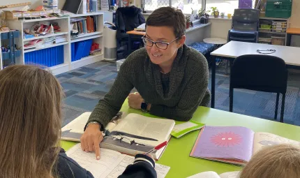 Teacher Ali Farmer is sitting in a classroom, at a table with two students pointing at a booklet of their school work.