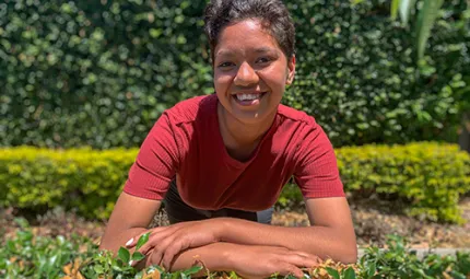 Young woman with short hair and red shirt leans on a hedge and faces the camera. In the background are more hedges.