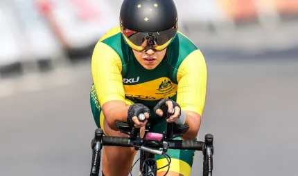 A woman dressed in green and yellow cycling wear rides a bike toward the camera. In the background is a dark road and signage.